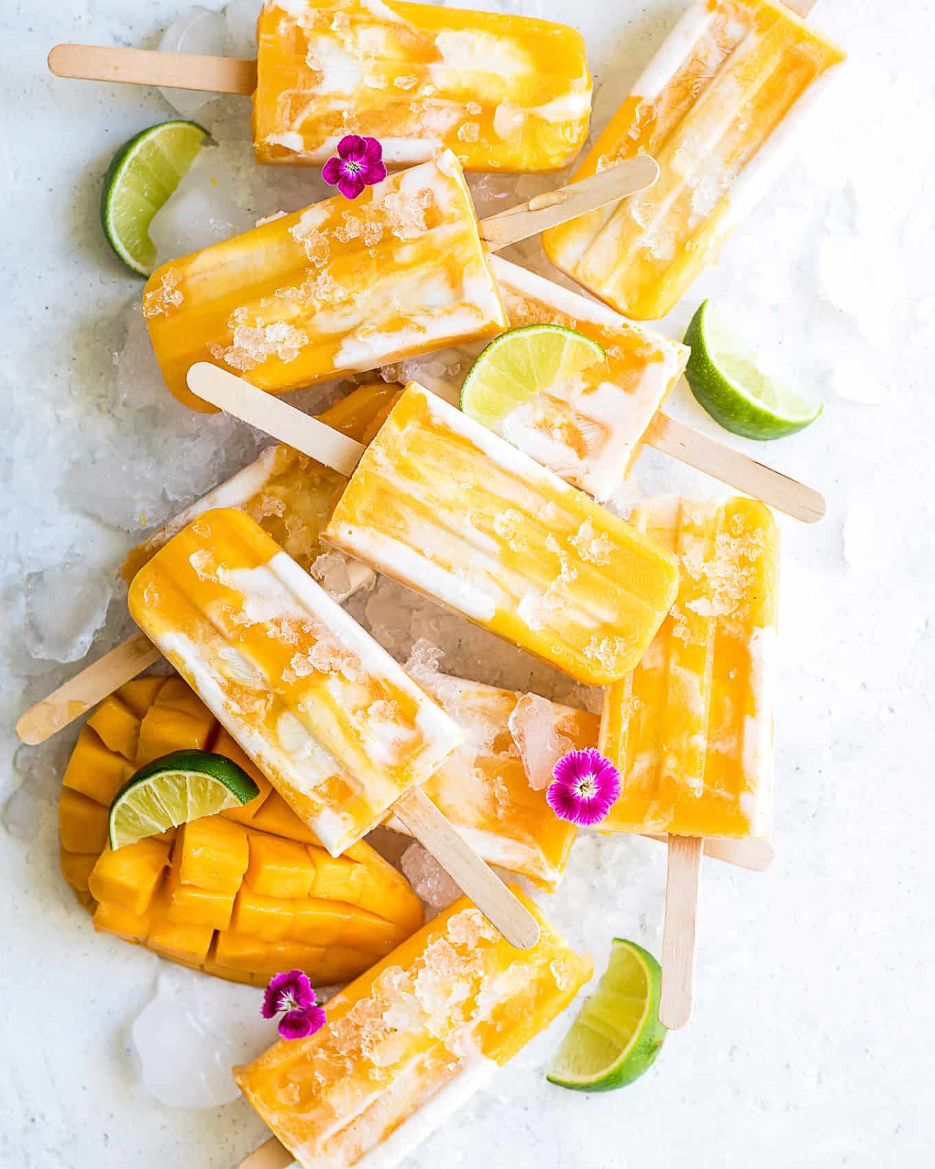 Mango popsicles on ice with mango and limes and pink flowers.