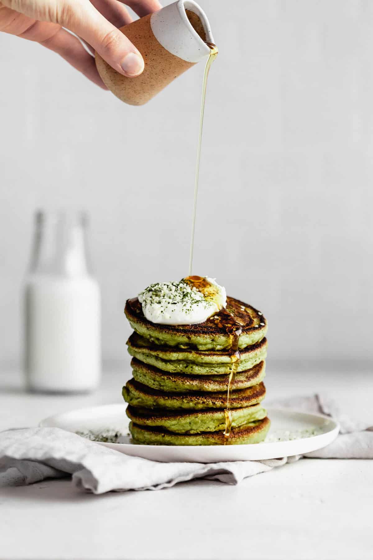 A hand pouring maple syrup on a stack of matcha pancakes.