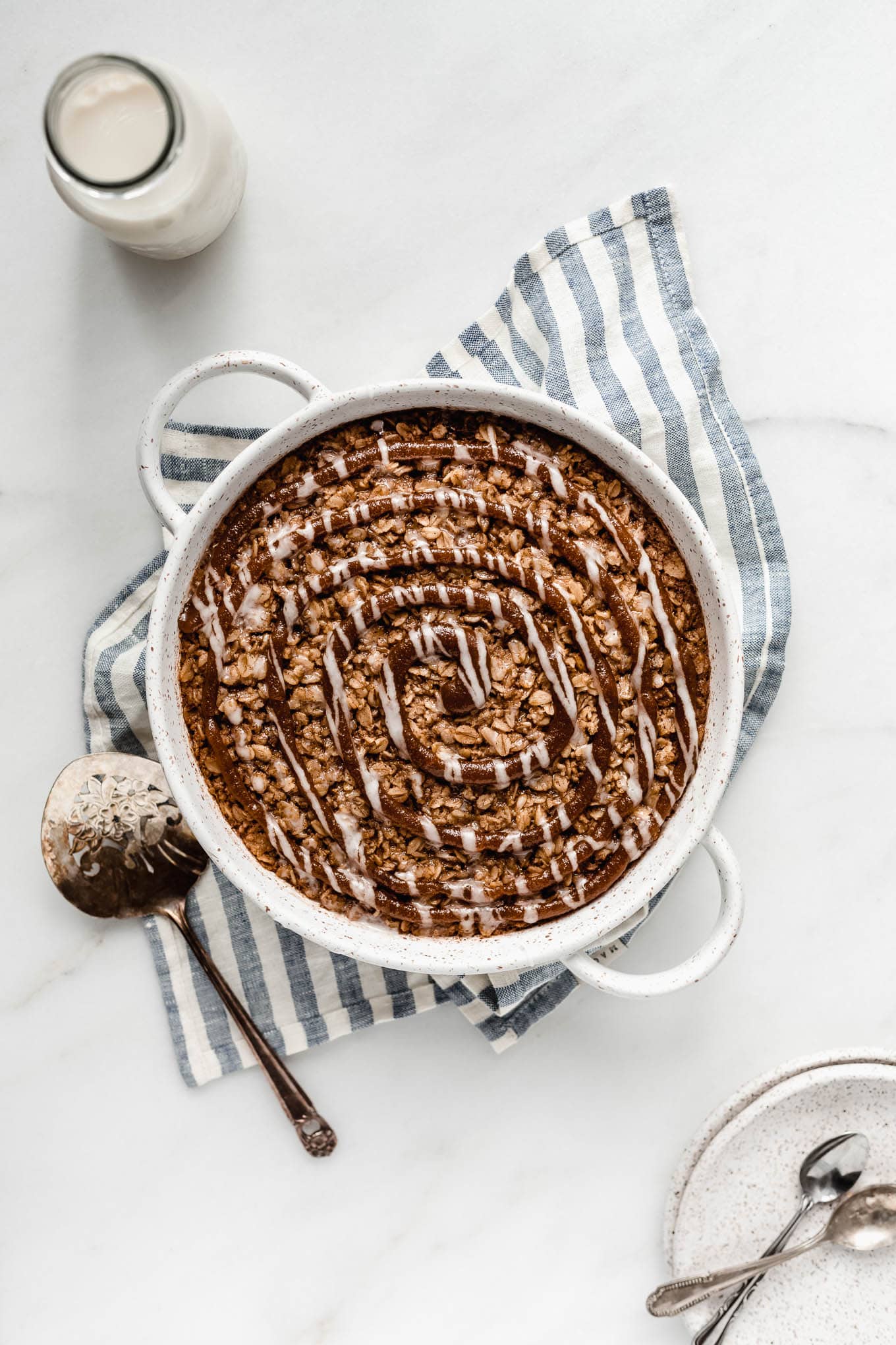 Cinnamon roll baked oats in a round baking dish on a blue and white stipe hand towel.