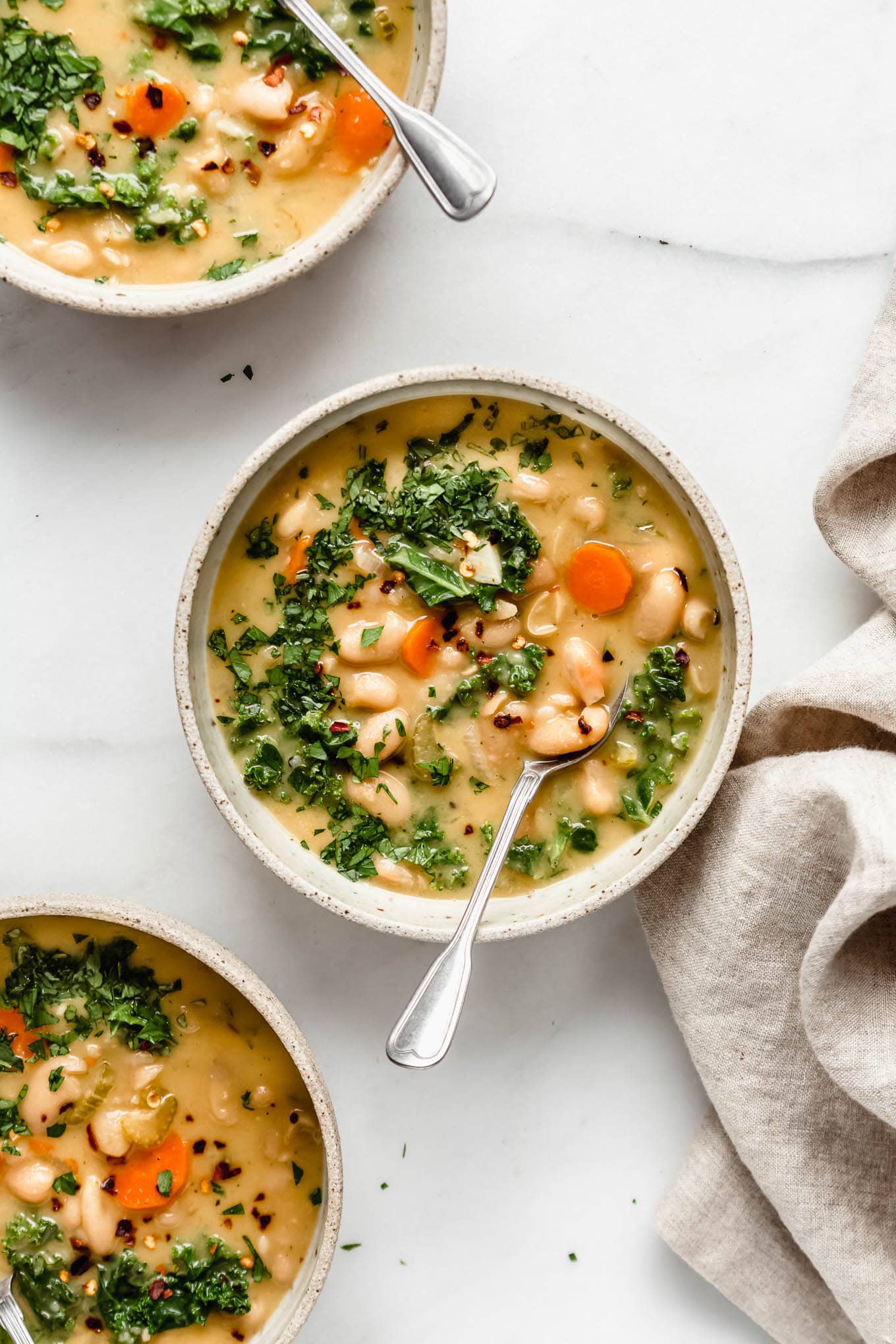 three bowls of tuscan white bean soup with a beige napkin on the side.