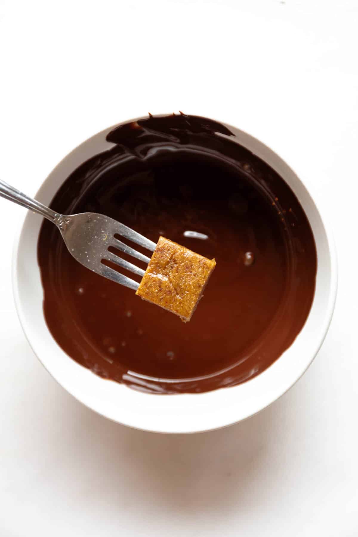 a fork holding a date caramel above a bowl of melted chocolate.