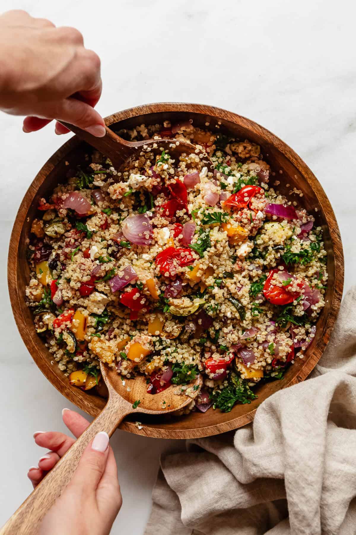 two hands scooping quinoa salad with roasted veggies out of a salad bowl.